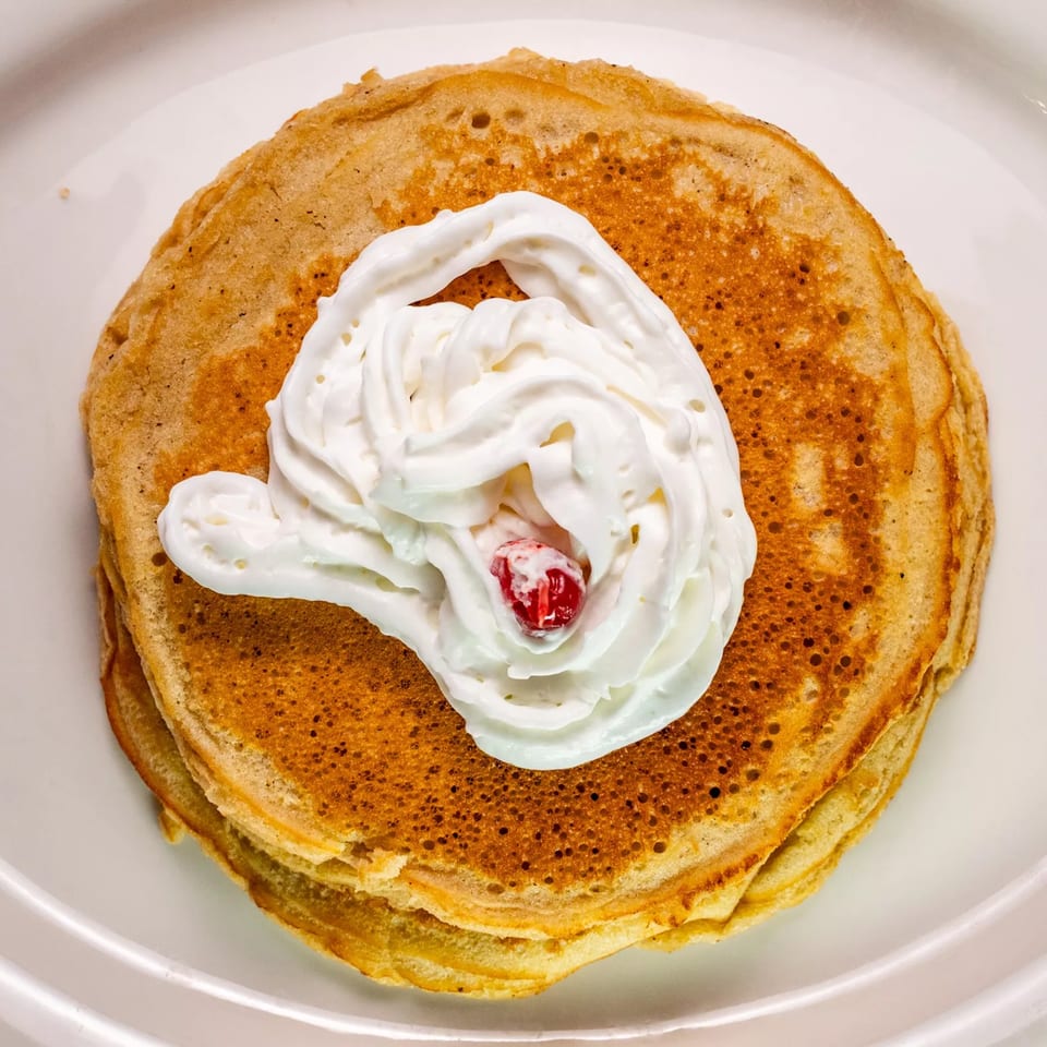 Stack of golden pancakes topped with whipped cream and a single red cherry on a white plate.
