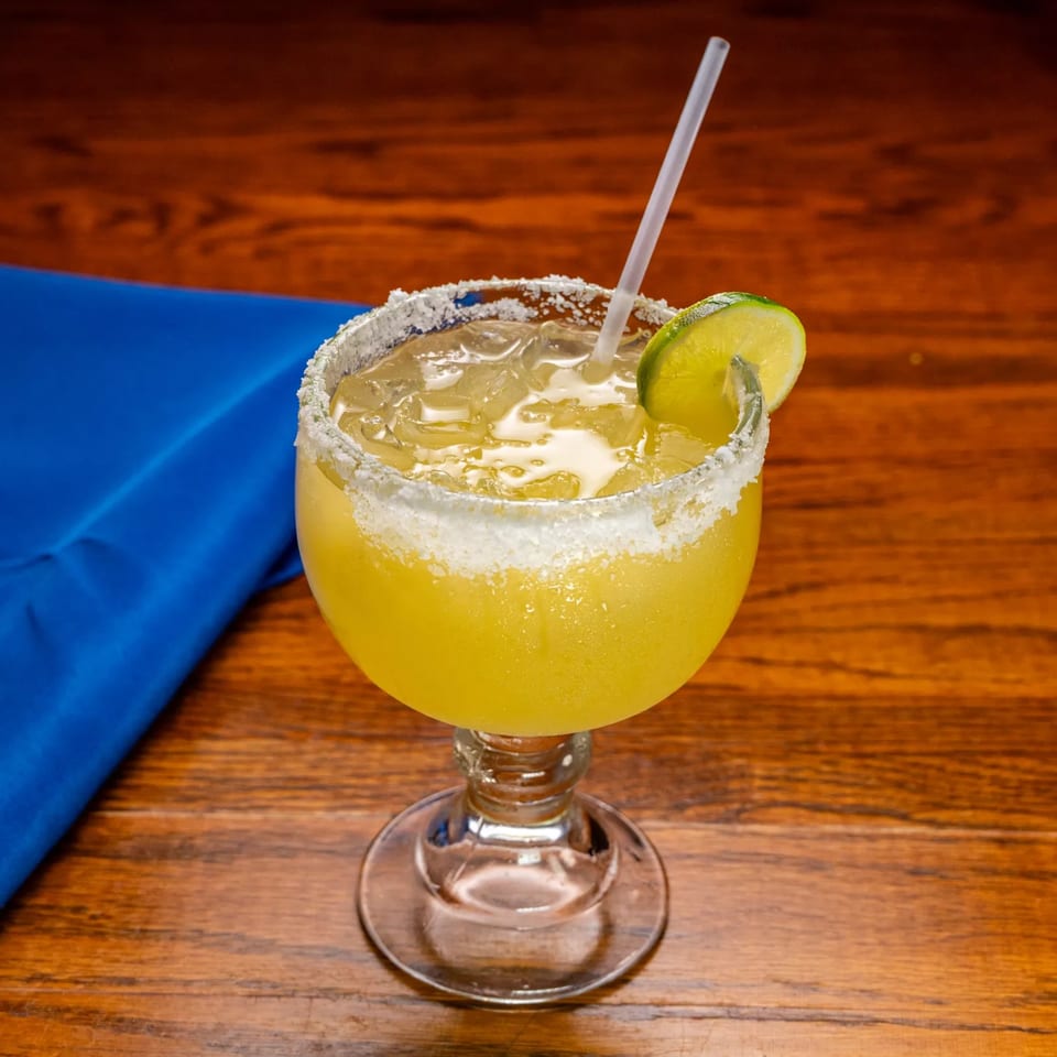 A salted margarita with a lime wedge and straw on a wooden table with a blue napkin.