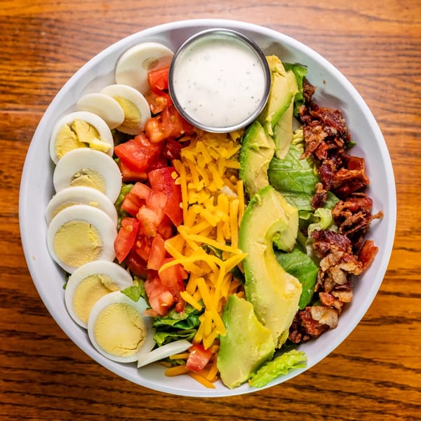 Overhead view of a Cobb salad with hard-boiled eggs, tomatoes, cheese, avocado, bacon, and ranch dressing.