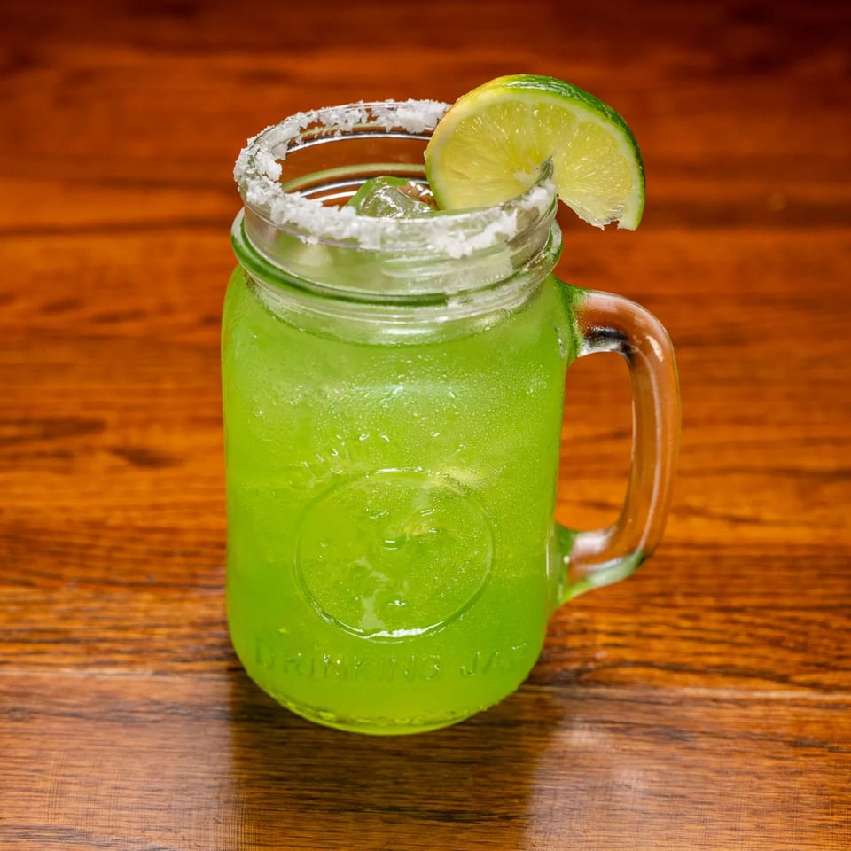 Bright green margarita in a mason jar with a salted rim and lime wedge on a wooden table.