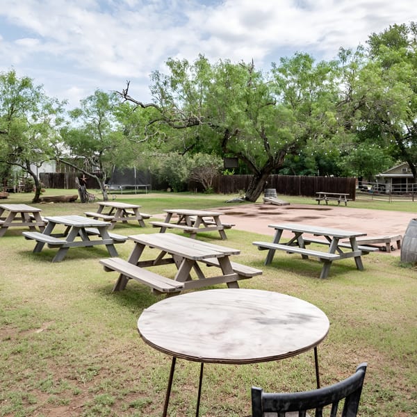 Outdoor dining area with multiple picnic tables on grass and dirt, surrounded by trees and a fence.