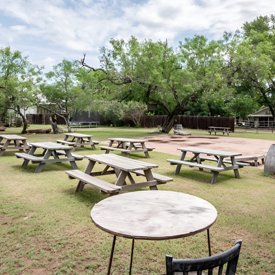 Outdoor dining area with multiple picnic tables on grass and dirt, surrounded by trees and a fence.