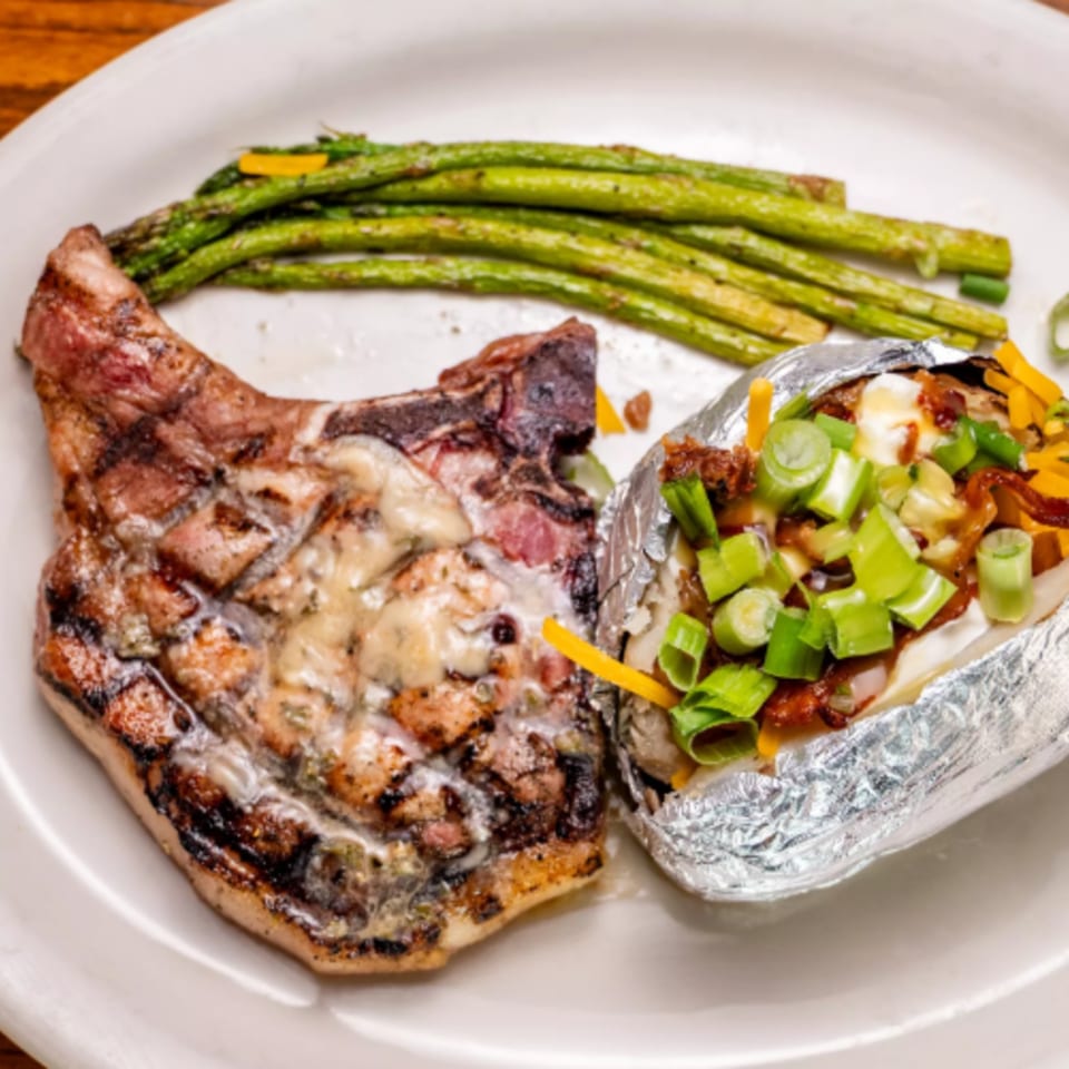 Grilled pork chop with melted cheese, a loaded baked potato, and asparagus on a white plate.