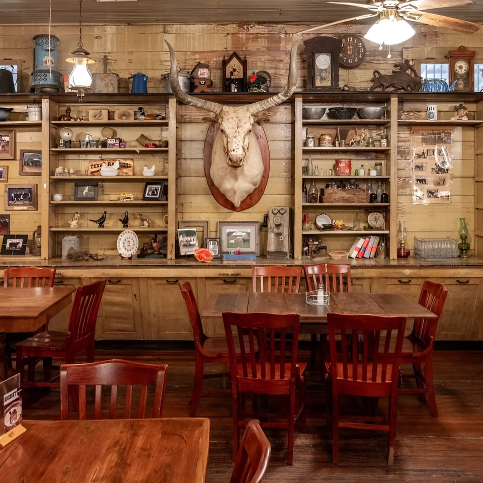 Rustic dining room with wooden tables and chairs, a mounted longhorn skull, and shelves filled with antique decor.