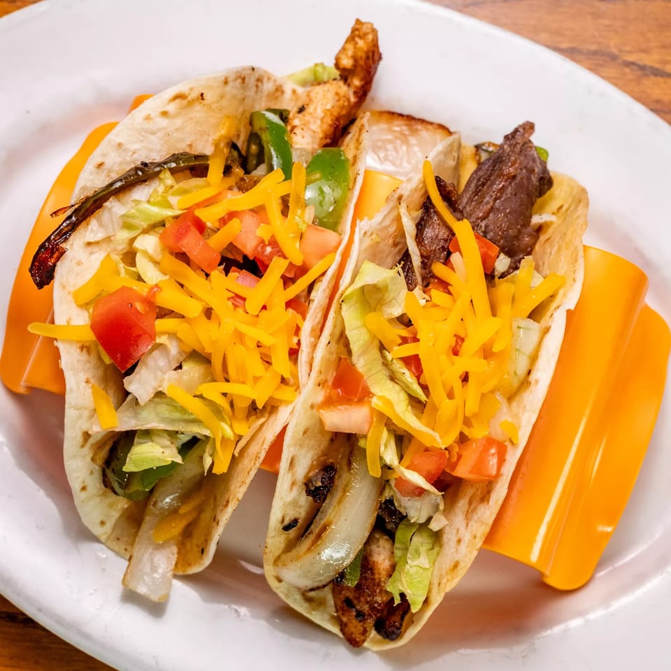 Two steak tacos with cheese, lettuce, and tomatoes, served on a white plate.