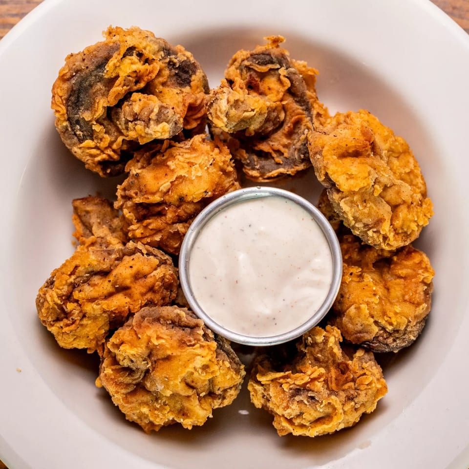 A white bowl filled with golden-brown fried mushrooms and a small cup of creamy dipping sauce.