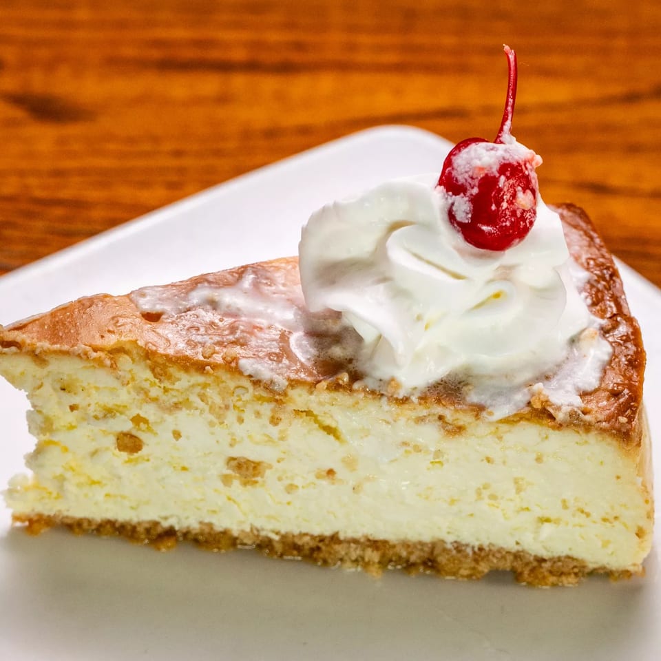 Slice of cheesecake with whipped cream and a cherry on top, on a white plate.