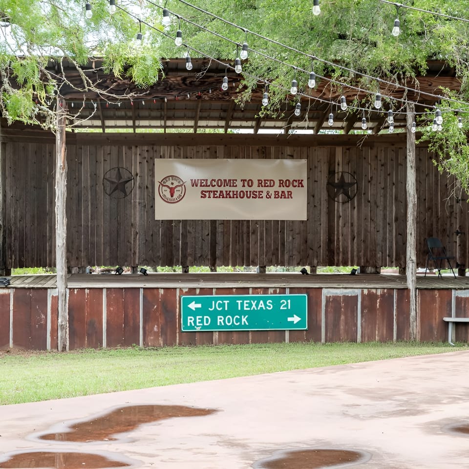 Outdoor stage with a "Welcome to Red Rock Steakhouse & Bar" sign, string lights, and a road sign.